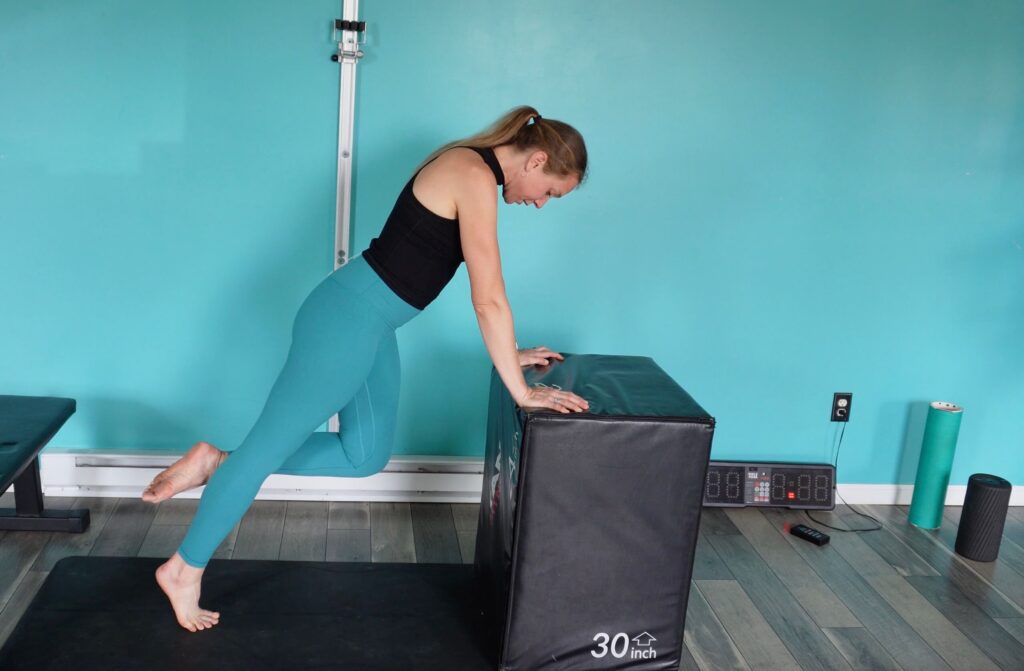 Dr. Chloe leaning forward supporting herself by her hands on a box with her hips extended forward standing on one foot. She is pressing through her toes to lift up onto the ball of her foot as a foot strengthening exercise for runners